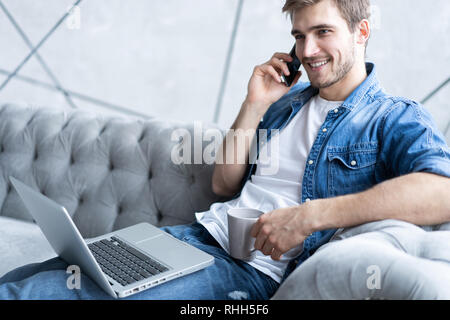 Portrait of handsome young man making a call and using his laptop while sitting on sofa at home Stock Photo