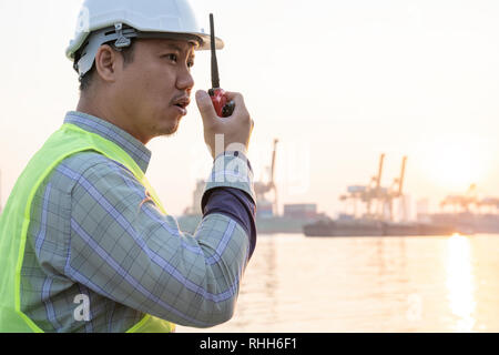 Asian harbor dock worker talking on radio with ship background - Image Stock Photo