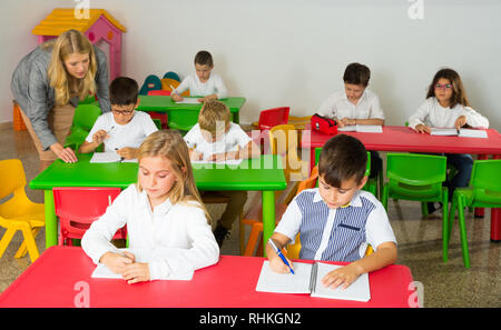 Schoolkids working at desks in classroom and teacher helping them Stock Photo