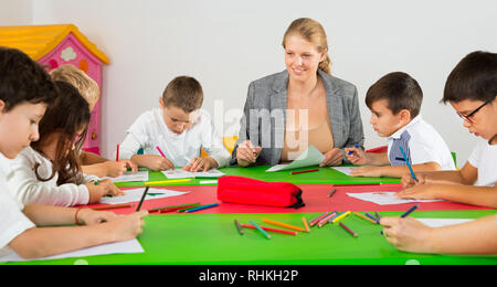 Friendly female teacher talking to children, sitting together around desk in a classroom Stock Photo
