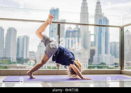 Happy little boy is practicing yoga on his balcony with a panoramic view of the big city Stock Photo