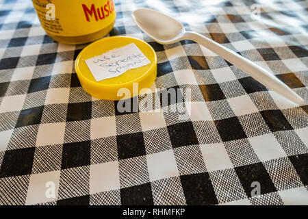Closeup of Colmans English Mustard jar with lid off and sticker saying use seperate spoon. plastic spoon on a black and white plastic gingham table cl Stock Photo