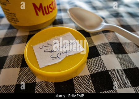 Closeup of Colmans English Mustard jar with lid off and sticker saying use seperate spoon. plastic spoon on a black and white plastic gingham table cl Stock Photo