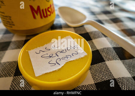 Closeup of Colmans English Mustard jar with lid off and sticker saying use seperate spoon. plastic spoon on a black and white plastic gingham table cl Stock Photo