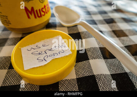 Closeup of Colmans English Mustard jar with lid off and sticker saying use seperate spoon. plastic spoon on a black and white plastic gingham table cl Stock Photo