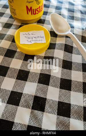 Closeup of Colmans English Mustard jar with lid off and sticker saying use seperate spoon. plastic spoon on a black and white plastic gingham table cl Stock Photo