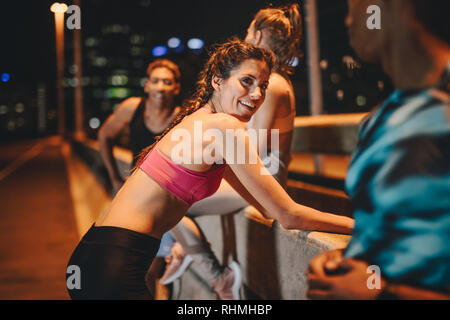 Group of men and women doing stretching and talking after workout session at night. Multi-ethnic friends taking break after workout at night. Stock Photo