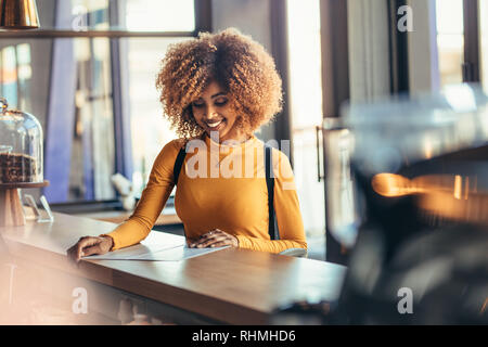 Smiling afro american woman standing at the billing counter of a coffee shop looking at the menu. Cheerful afro american traveller ordering food at a Stock Photo