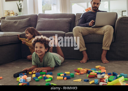 Man working on laptop sitting at home with kids playing on the floor. Cheerful kids playing with building blocks and watching television at home. Stock Photo