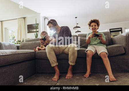 Happy man having fun with his kids playing video game at home. Father playing video game sitting on couch with his two kids. Stock Photo