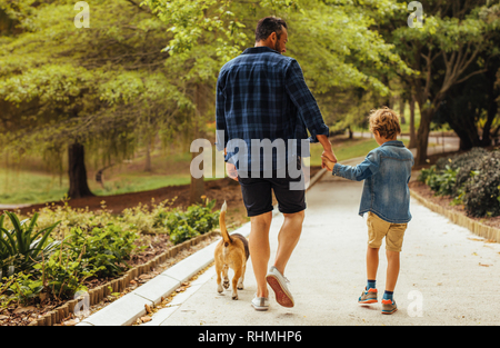 Rear view of father and son with dog walking in a park. Man holding hand on his little boy walking and talking outdoors in park. Stock Photo