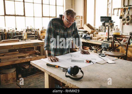 Senior carpenter studying drawing before starting his work at workshop.  Carpenter checking drawing in carpentry workshop. Stock Photo
