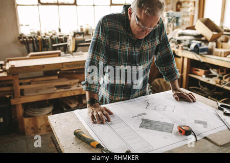 Senior carpenter reading drawing in workshop bench. Mature man studying building floor plan before starting work at carpentry workshop. Stock Photo