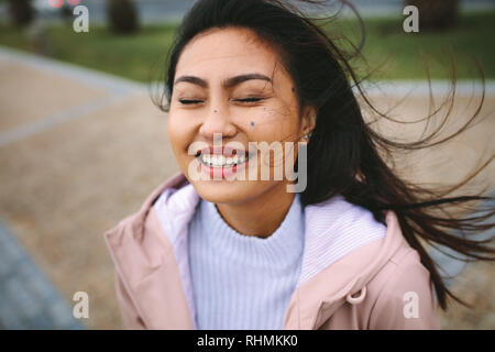 Smiling woman standing outdoors enjoying the breeze with her eyes closed. Portrait of a woman standing outdoors with her hair flying. Stock Photo