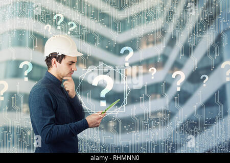 Puzzled engineer wearing protective helmet using a tablet computer gadget looking thoughtful to get answer to many questions. Science searching and in Stock Photo