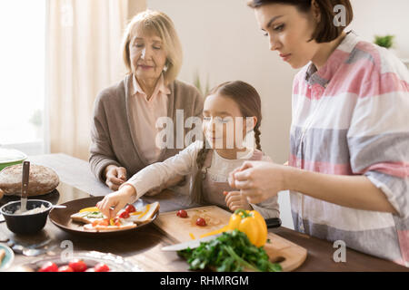 Little girl putting pieces of fresh vegetables on toast while helping her mom and granny with sandwiches for breakfast Stock Photo