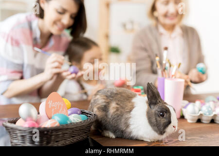 Cute fluffy rabbit sitting on wooden table by basket with Easter eggs being painted for holiday by two women and little girl Stock Photo
