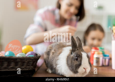 Adorable rabbit and heap of Easter eggs on table on background of mother and daughter painting Stock Photo