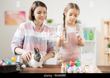Adorable girl painting Easter egg in silver color with her mom standing near by and cuddling fluffy rabbit Stock Photo