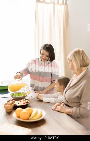 Young woman pouring fresh orange juice from jug into glass by served table during breakfast with her mom and little daughter Stock Photo