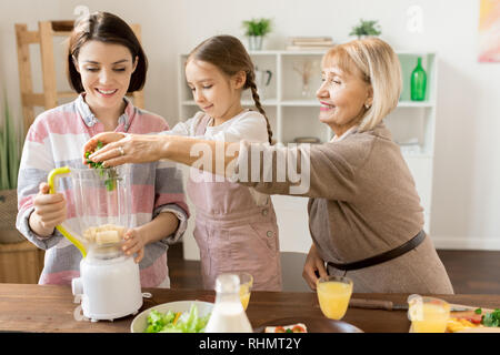 Mature woman and her granddaughter putting fresh green vegetables into blender while preparing smoothie for all family Stock Photo