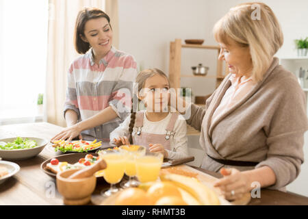 Kind grandma cuddling her granddaughter while cooking breakfast by table in the kitchen Stock Photo