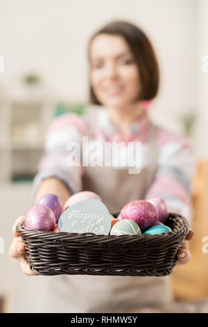 Heap of colorful painted Easter eggs and small greeting card in basket held by young casual woman Stock Photo