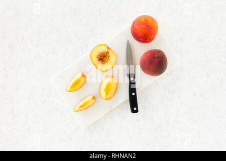 Whole and cut peaches and a knife on marble chopping board and white, gray, textured and patterned background. Top view. Stock Photo