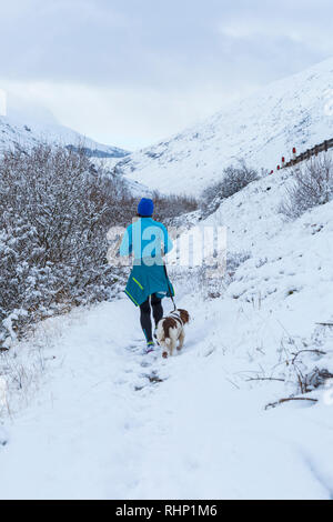 Woman running along snow covered path with dog at Glencoe, Highlands, Scotland in winter - rear back view Stock Photo