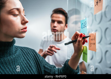Team of project managers having meeting at modern office. Business colleagues discussing new ideas and using sticky glass note wall Stock Photo