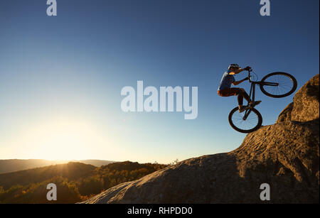Cyclist standing on back wheel on trial bike. Professional sportsman rider making acrobatic stunt on the edge of big boulder on the top of mountain at sunset. Concept of extreme sport active lifestyle Stock Photo
