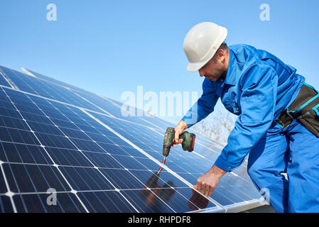 Man worker in blue suit and protective helmet installing solar photovoltaic panel system using screwdriver. Professional electrician mounting blue solar module. Alternative energy ecological concept. Stock Photo
