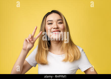 Pleased asian woman in t-shirt showing peace gestures and looking at the camera over yellow background Stock Photo