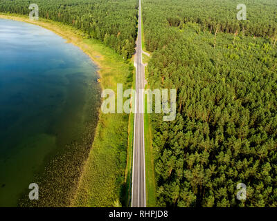 Aerial top down view of two-lane road with pine tree forests on one side, and deep green lake on the other. Beautiful summer scenery in Lithuania. Stock Photo