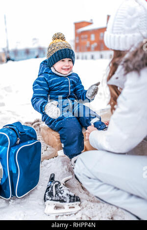 Young mother wears socks for skates, helps a little boy 3-5 years old child. In winter, on the rink in the city. Rest on fresh frosty air on skates Stock Photo