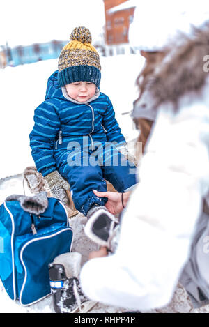 A young mother puts on skates, helps a little boy of 2-4 years old child. In winter, on the rink in city. Rest on the fresh frosty air on skates Stock Photo