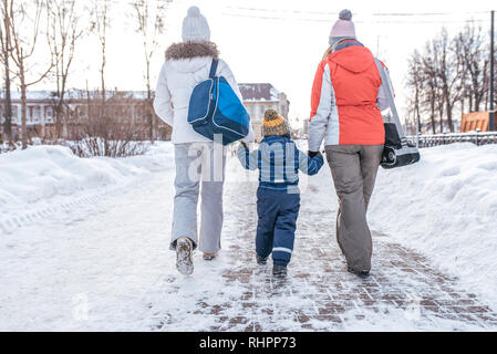 Mom and girlfriend sees the boy s son 3-5 years old by hand walking down the street. Back view. Winter in city on background of snowdrifts. Returning Stock Photo