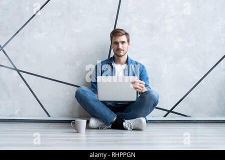 Young man working on laptop sitting on floor. Guy with computer reading news, surfing net or freelancing. Stock Photo