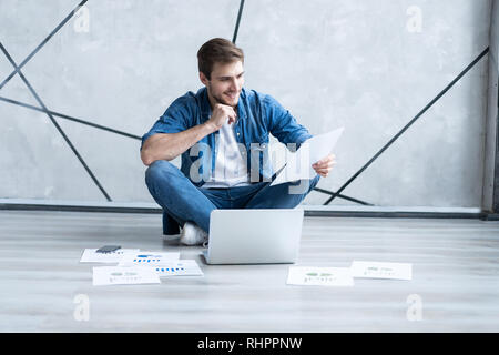 Man working at home. Handsome young man sitting on the floor and examining document while laptop and documents laying near him. Stock Photo