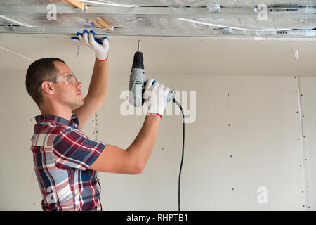 Young man in goggles fixing drywall suspended ceiling to metal frame using electrical screwdriver on ceiling insulated with shiny aluminum foil. Renov Stock Photo