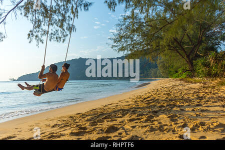 A young couple in beach wear swing on a basic swing over a golden sand beach in a paradise location. Stock Photo