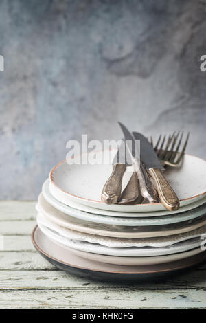 Stack of clean dinner plates with cutlery on top over wooden table. Pastel tones. Stock Photo