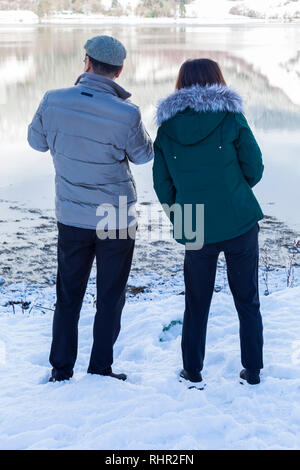 Couple admiring the views reflected in Loch Leven and snow on a cold winters day at Glencoe Village, Highlands, Scotland in winter Stock Photo