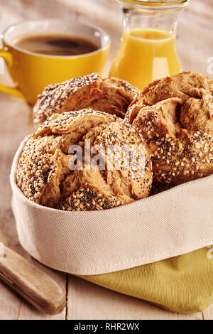 Fresh baked dark bread buns with seeds, served in fabric bag with juice and coffee in background on wooden table Stock Photo