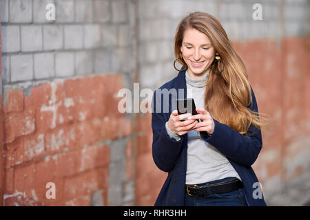 Beautiful blonde girl wearing a blue coat walking in front of a brick wall while typing on her smart phone