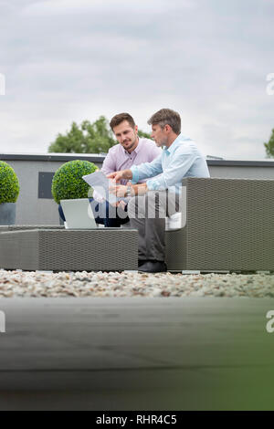 Businessmen discussing over documents with laptop while sitting at office terrace Stock Photo