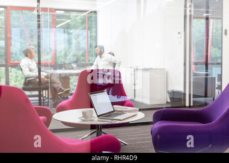 Laptop on table by chairs in boardroom with businessmen discussing in background Stock Photo