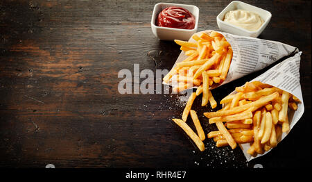 Paper cones with salted French fries spilling out onto dark wood with copy space served with sauces Stock Photo