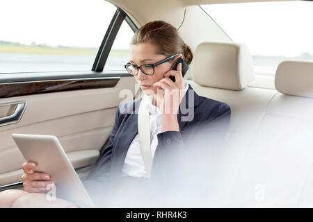Businesswoman talking on smartphone while using digital tablet in car Stock Photo