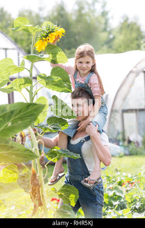Smiling mother carrying daughter on shoulders by sunflower plant at farm Stock Photo